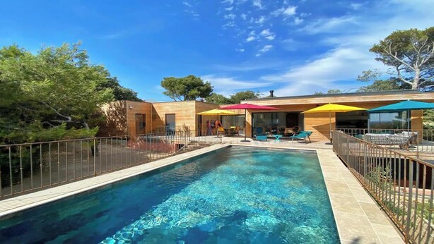 Une piscine avec une terrasse en bois, entourée d’une clôture, et un bâtiment agrémenté de parasols colorés et doté d’un espace de détente extérieur.