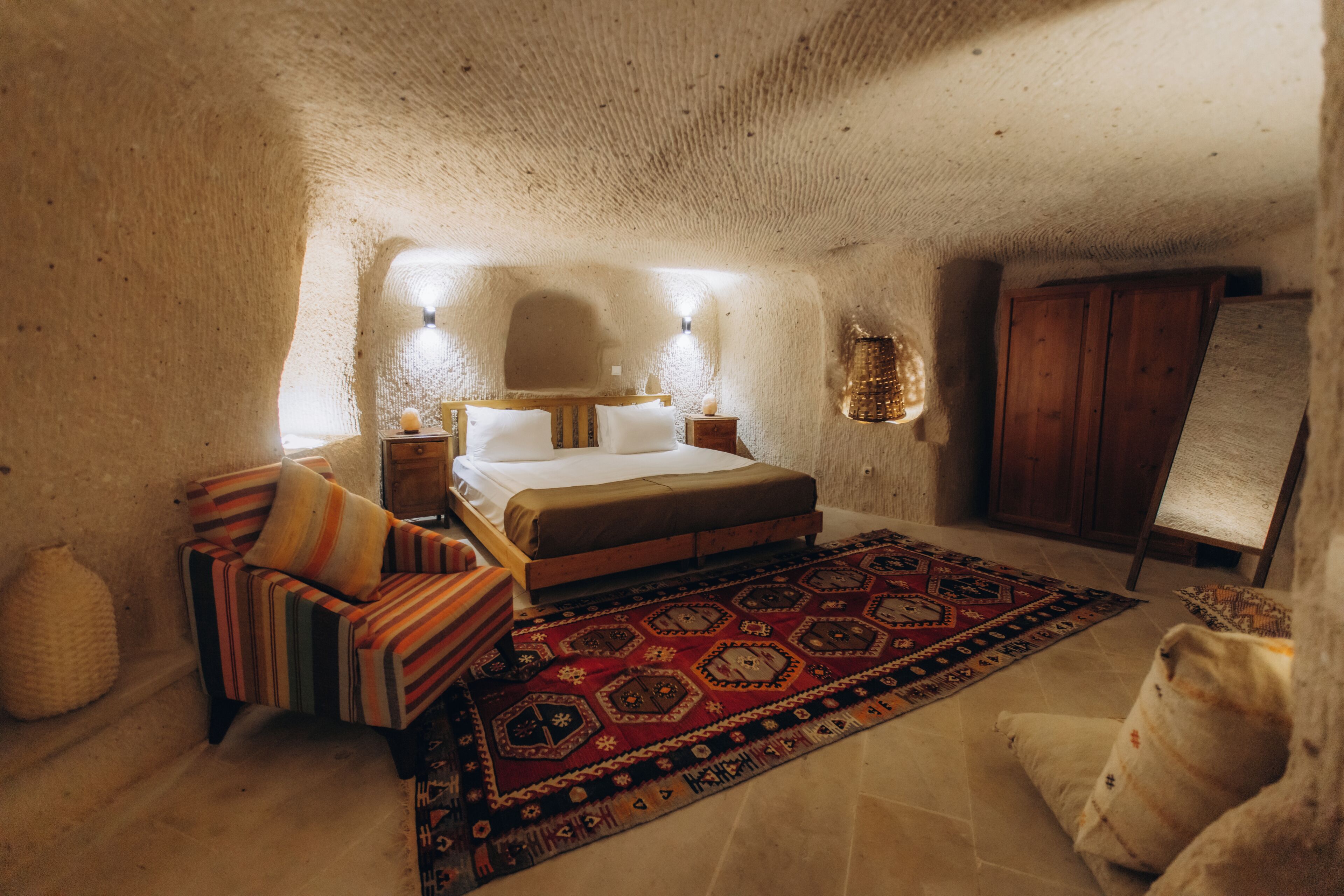 A cozy bedroom with a bed, pillows, a striped chair, a patterned rug, and a wooden cabinet.