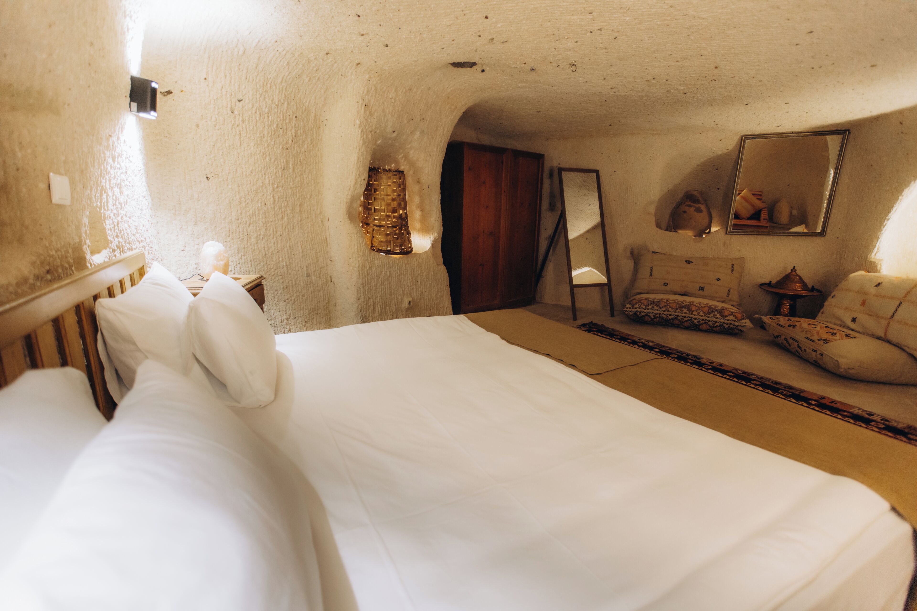 A room with a large bed, a wooden headboard, a small table with a lamp, a mirror, and a sofa with patterned cushions.