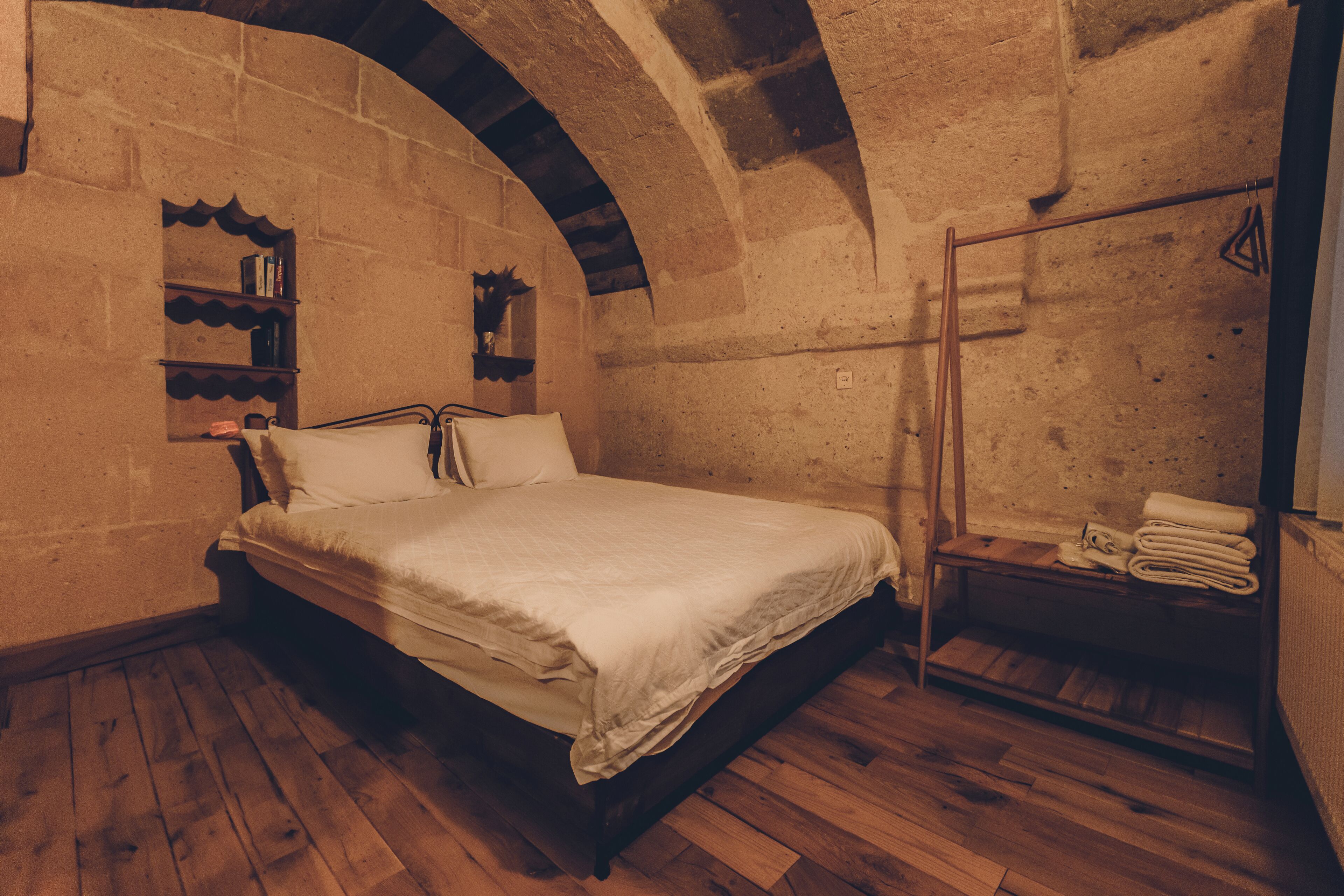 Une chambre avec un plancher en bois, un lit recouvert d’une courtepointe blanche, un banc en bois avec des serviettes, et des murs en pierre avec un plafond en bois.