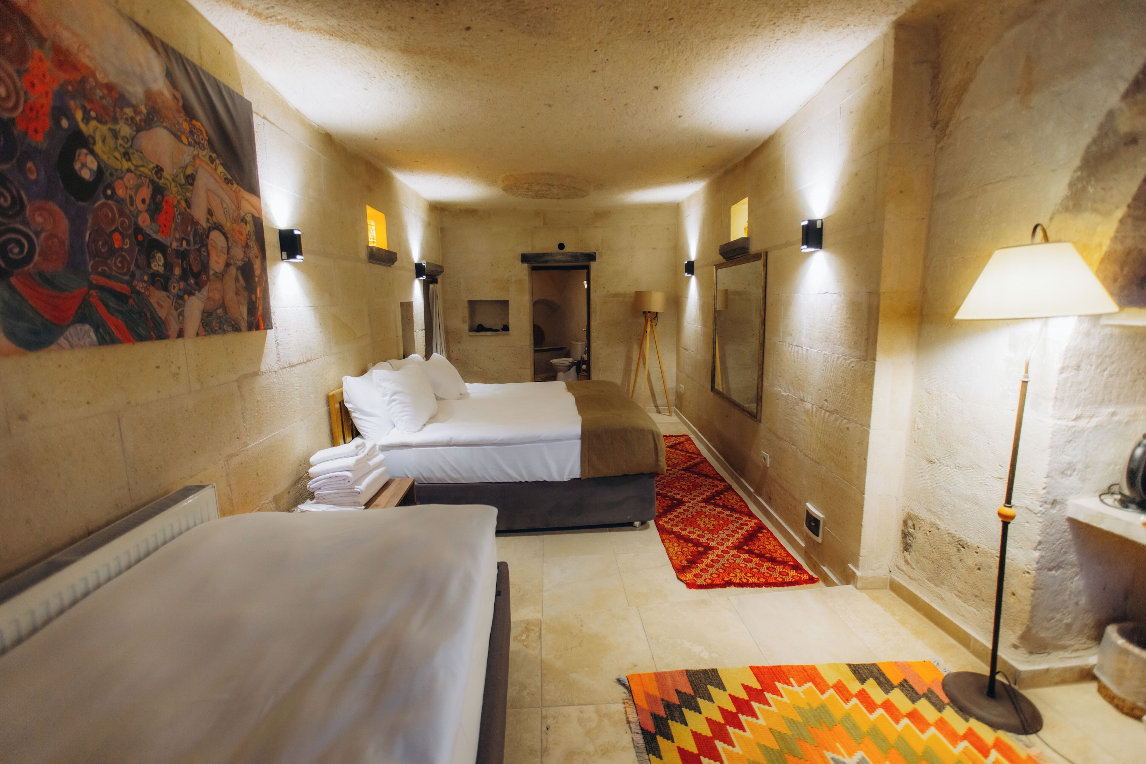 A hotel room with two beds, a red patterned rug, a standing lamp, and a painting on the wall.