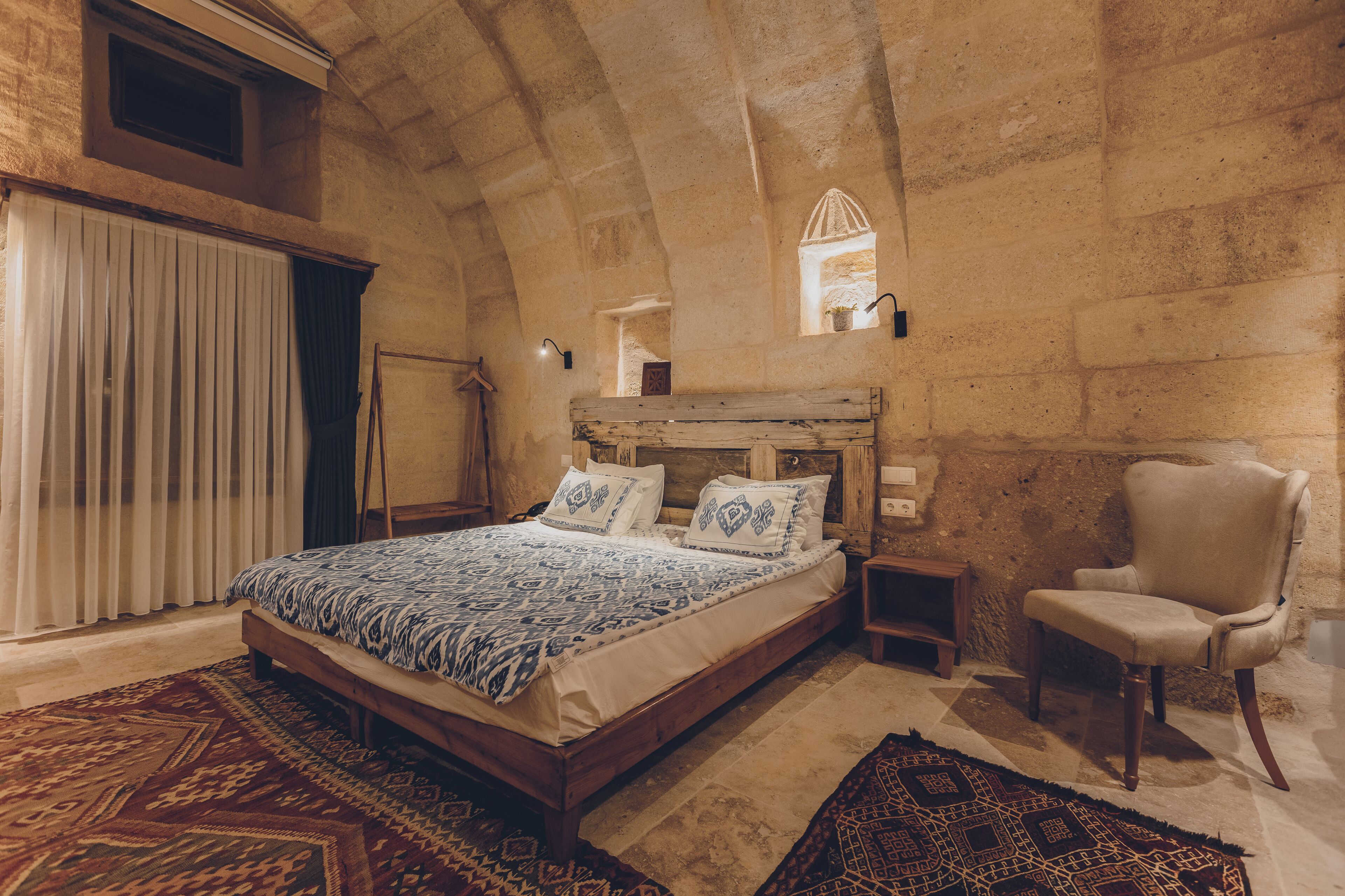 A bedroom with a wooden bed, a chair, a nightstand, and a stone wall.