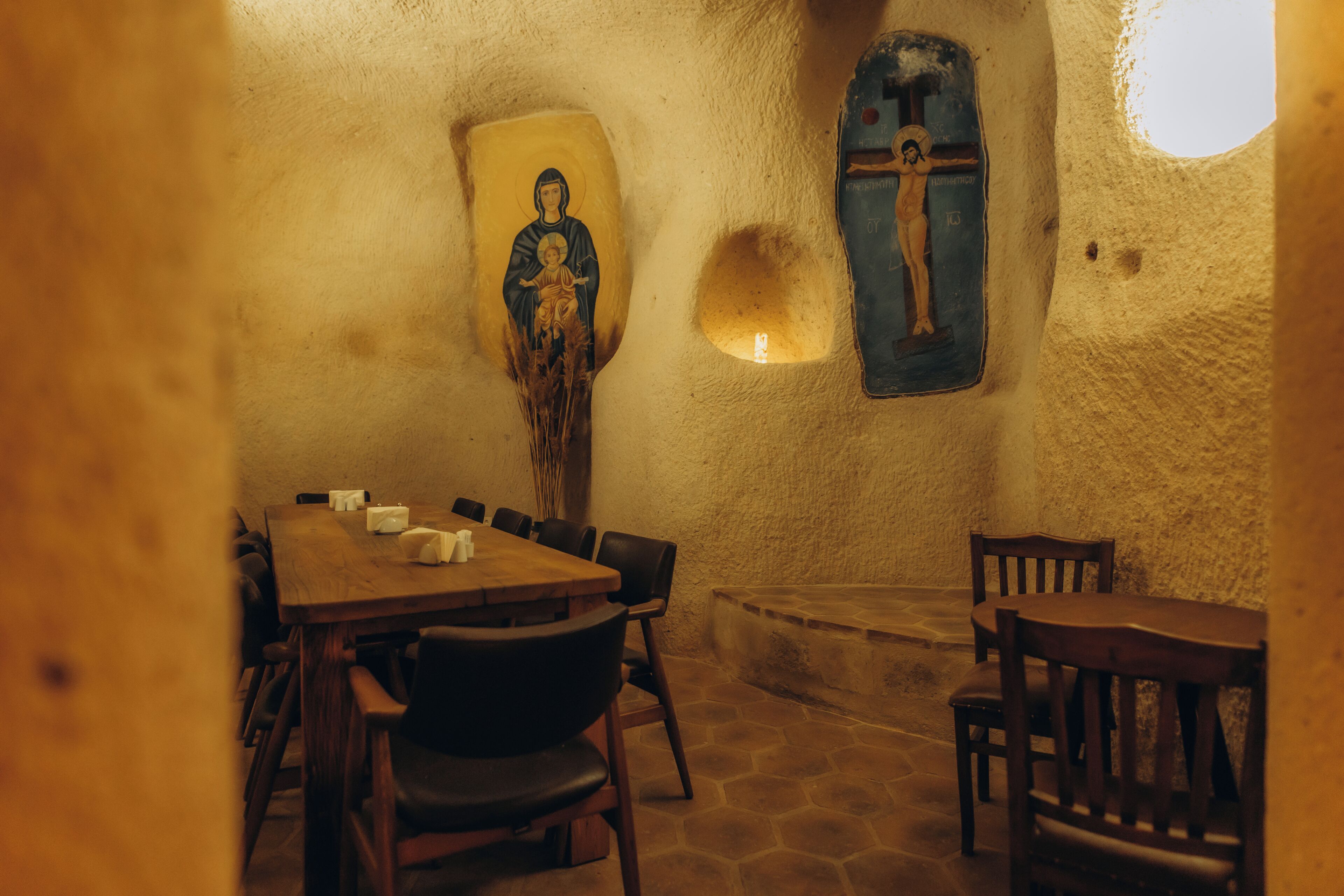 A dining area with a wooden table and chairs, a stone floor, and religious icons on the walls.