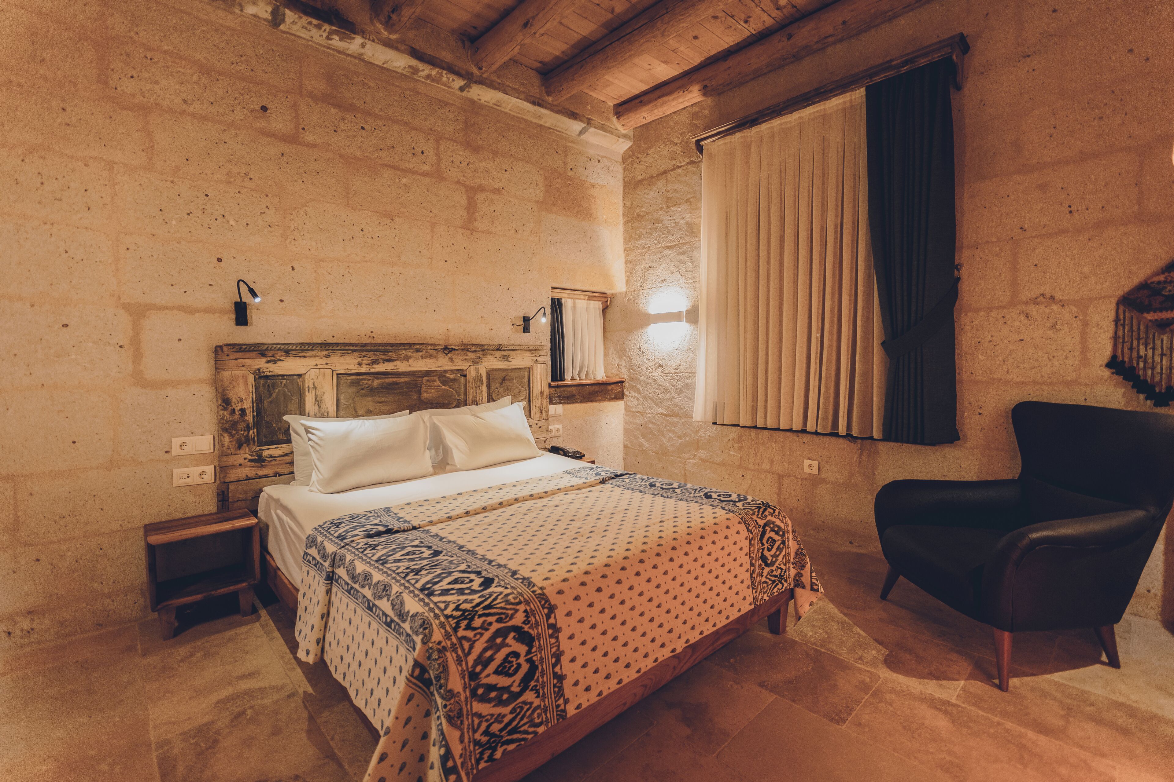A bedroom with a wooden headboard, a bed with white linens, a patterned bedspread, a black leather chair, and a wall-mounted shelf with a lamp.