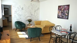 Una sala de estar con un sofá amarillo, sillones verdes, una mesa de comedor con sillas, un cuadro colgado en la pared y una lámpara de techo.