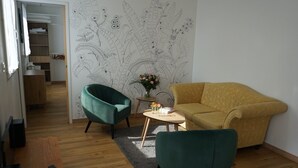 Un salón con un sofá amarillo, un sillón verde y una pequeña mesa con un florero. Se ve un suelo de madera y una escalera al fondo.