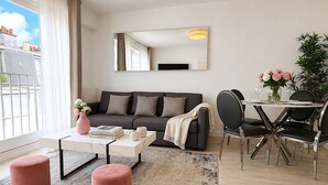 A modern living room with a grey sofa, a coffee table, two pink ottomans, a dining area with a table and chairs, and a large mirror on the wall. - L'appartement Bouquet  (Paris)