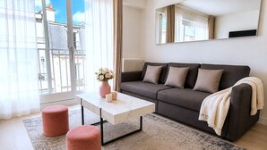 A modern living room with a grey sofa, white coffee table, pink ottomans, and a large window with curtains. - L'appartement Bouquet  (Paris)