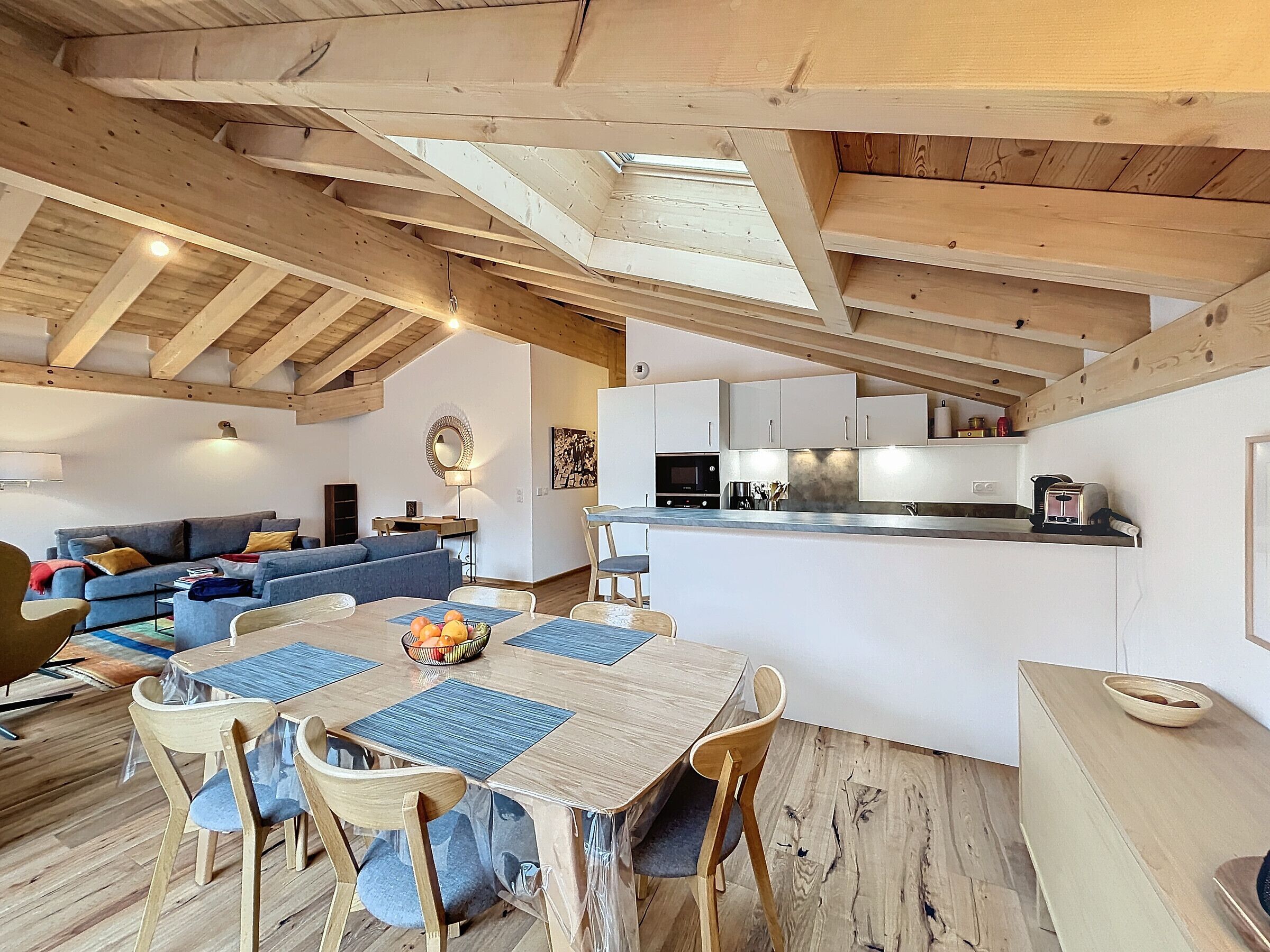 Ein geräumiges Loft mit hoher Decke, freiliegenden Holzbalken, einem Essbereich mit Tisch und Stühlen, einer Küche mit weißen Schränken sowie einem Wohnbereich mit Sofa und Fernseher.