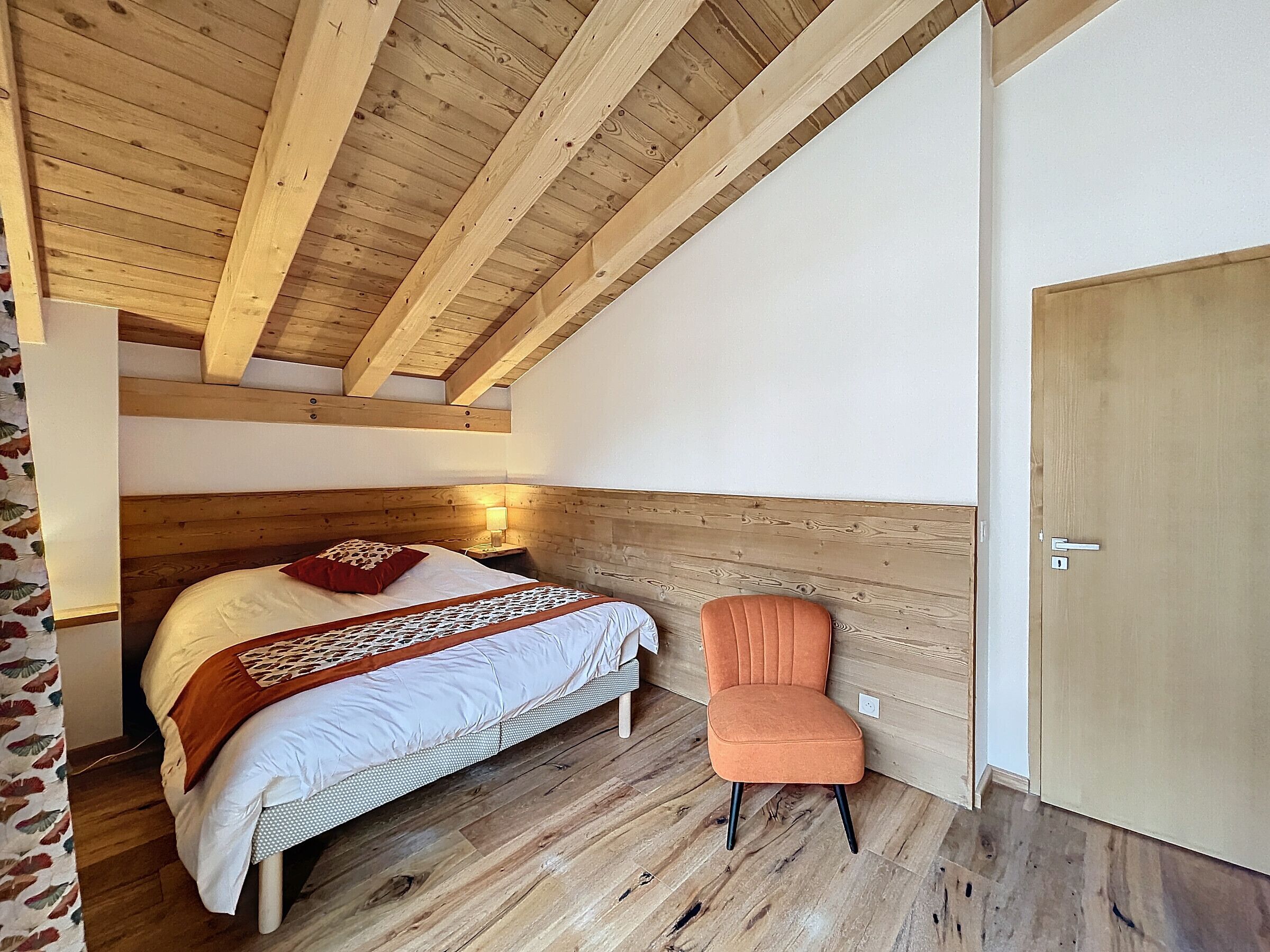 A bedroom with a wooden ceiling, a bed with a red pillow, a chair, and a wooden wardrobe.