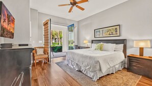 Ein modernes Schlafzimmer mit einem großen Bett, einem Deckenventilator, einem Schreibtisch mit Lampe und Blick auf einen Garten durch eine offene Tür.