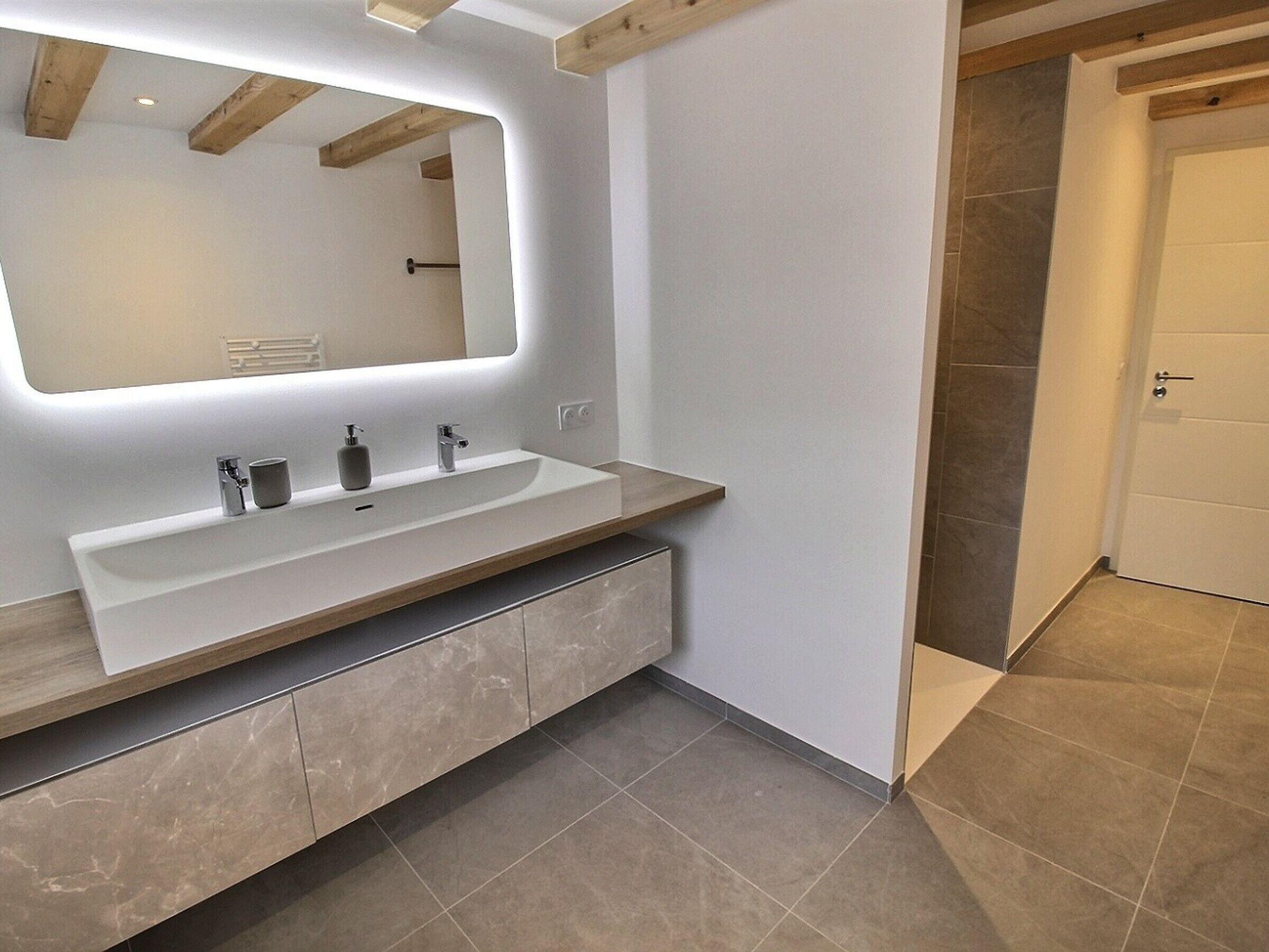 A modern bathroom with a large mirror, a white sink, and a wooden vanity.
