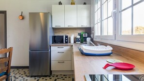 A compact kitchen with a stainless steel refrigerator, white cabinets, a microwave, and a coffee maker. There's a window providing natural light and a checkered floor pattern. - Single-story house with garden and parking in Yvetot-Bocage (Yvetot-Bocage)