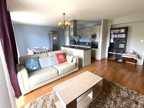 A living room with a sofa, coffee table, and a kitchen area with cabinets and appliances. - Centrally Located Perros-Guirec Apartment with WiFi, Balconies, and Parking (Perros-Guirec)