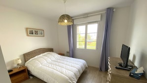 Ein Schlafzimmer mit einem großen Bett, einem Nachttisch mit Lampe, einem Schreibtisch mit Computer und Fernseher sowie einem Fenster mit Vorhängen.