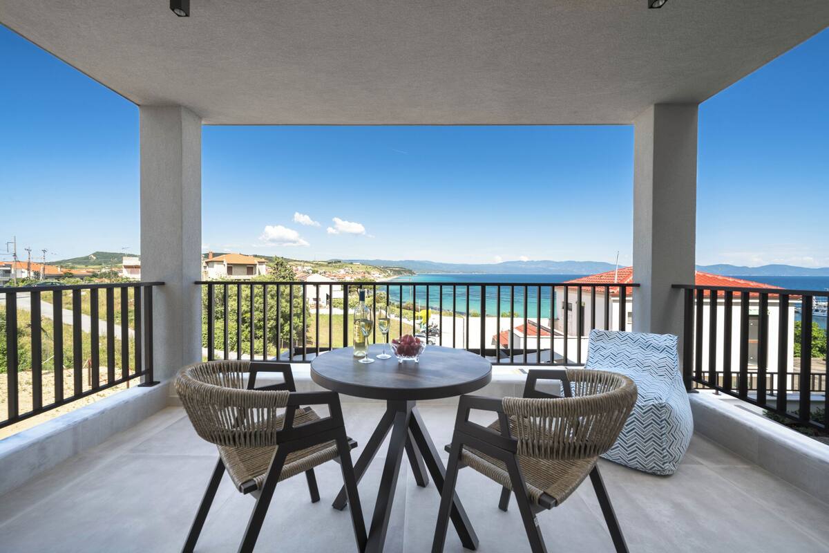 Een balkon met een ronde tafel en vier rieten stoelen, met uitzicht op de kustlijn met huizen en een strand.