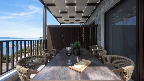Une table à manger entourée de chaises en osier, un verre de vin rouge et une planche de fromages, le tout sur un balcon offrant une vue sur la mer.
