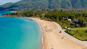 Een strand met helder turquoise water, een zandstrand en een achtergrond van groene heuvels.