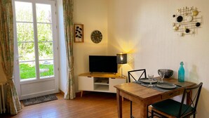 Ein gemütliches Zimmer mit Holzboden, einem Fernseher, einer Lampe, einem Esstisch mit Stühlen und einem Fenster mit Vorhängen.