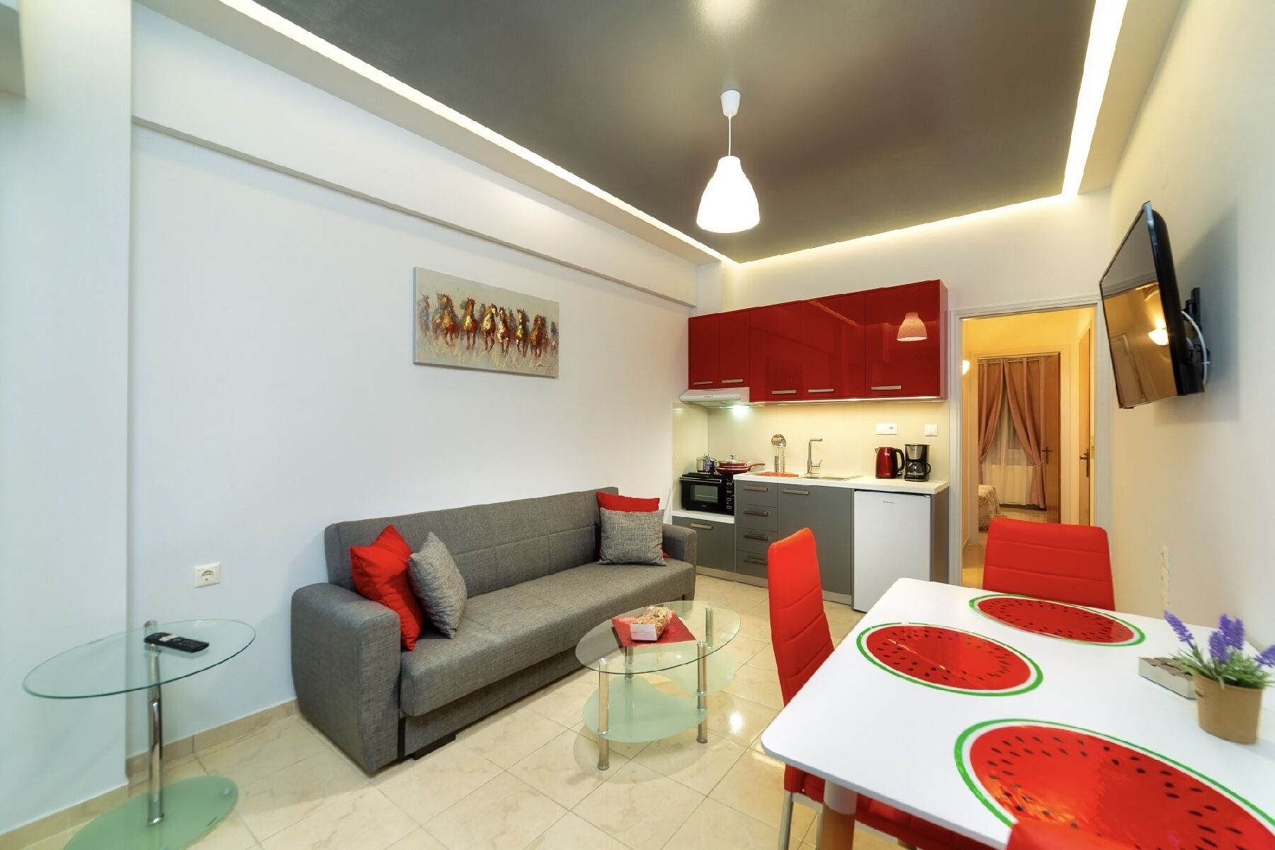 A compact living space with a grey sofa, a glass coffee table, a red dining table with a watermelon design, and a kitchen area with red cabinets and a TV mounted on the wall.
