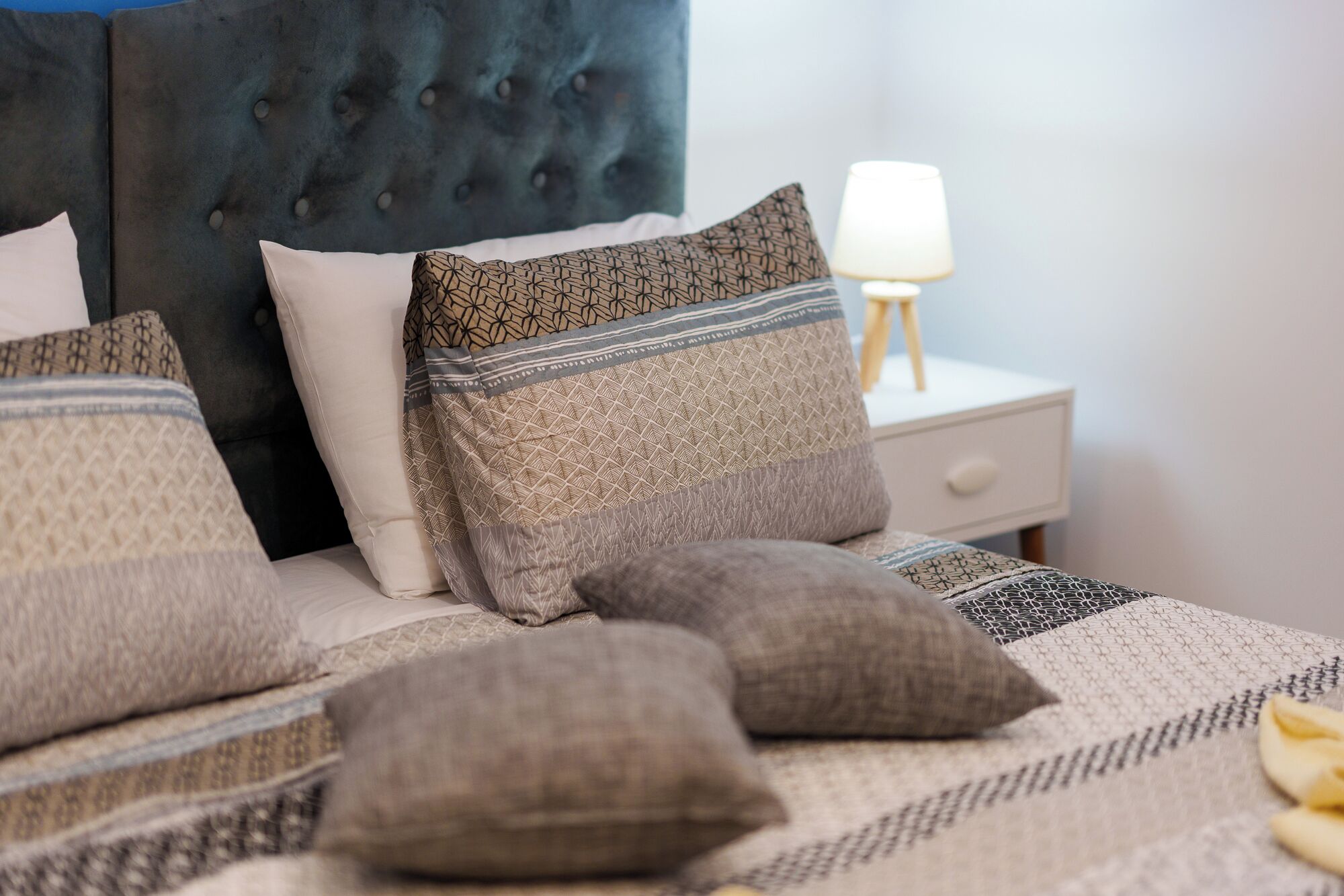 Ein ordentlich bezogenes Bett mit gemusterten Betttextilien und Kissen, ein gebürstetes Kopfstück und ein Nachttisch mit einer Lampe.