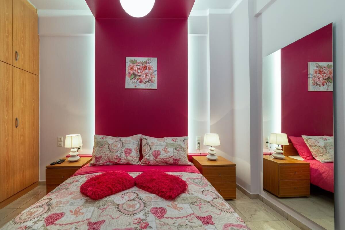 Een slaapkamer met een rode wand, een bloemenbedekker, twee bedlampen, een spiegel en een houten garderobe.