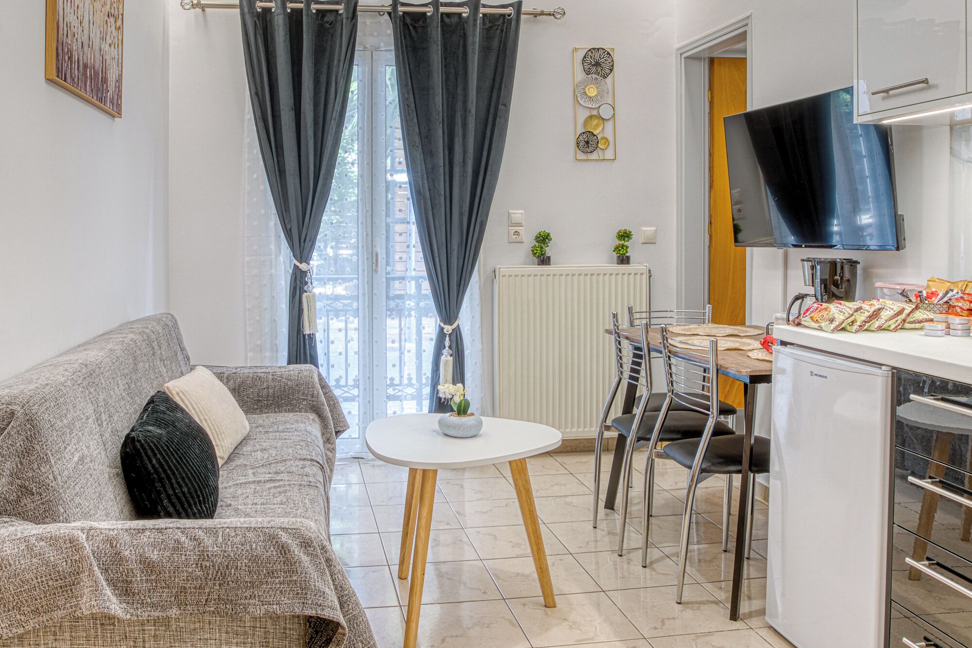 Ein modernes Wohnzimmer mit grauem Sofa, einem kleinen weißen Tisch und einer Küchenzeile inklusive Fernseher, Kühlschrank und Esstisch mit Stühlen.