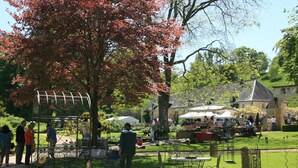 An outdoor market with tables and chairs, surrounded by greenery and a stone building in the background.