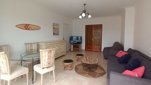 A living room with a glass-top dining table, two white chairs, a sofa with colorful cushions, a television, a wooden cabinet, and a door with a decorative glass panel. - Spacieux et Lumineux T2 au Cœur de Biarritz Proche e la Plage + Parking Privatif (Biarritz)