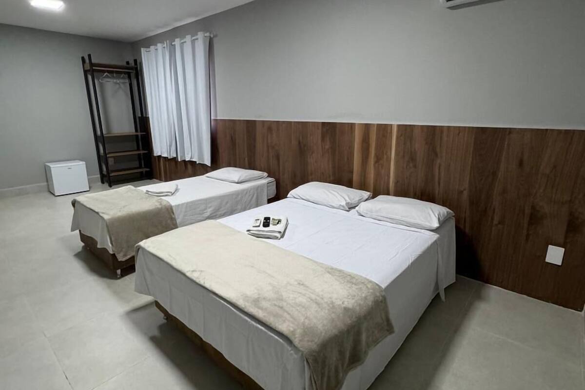 Une chambre d’hôtel avec deux lits simples, une tête de lit en bois, un appareil de climatisation, un bureau avec une chaise, une armoire et une fenêtre avec des rideaux.