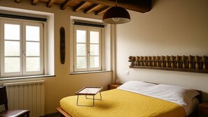 A bedroom with a bed, a chair, a radiator, two windows, a shelf with decorative items, and a pendant light.