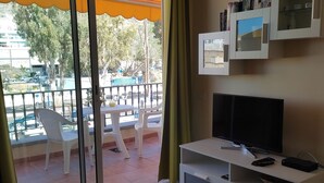 Un salon équipé d’un téléviseur, d’un meuble de rangement blanc, d’une table ronde avec un magazine, et d’un balcon avec des chaises et une table.