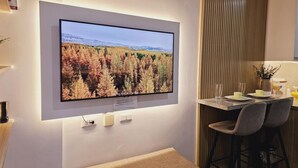 Ett modernt kök med en stor platt-TV som är monterad på väggen, ett matbord i trä med stolar och en liten bardisk med barstolar.