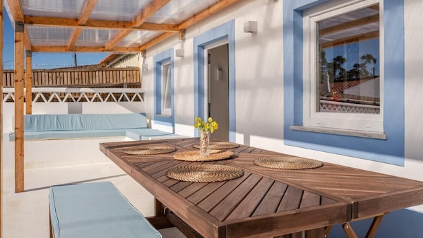 An outdoor patio with a wooden table, blue cushions, and a view of the sea. - Cottage in Aljezur- 2 bed Ensuite-casinha da Ivone (Aljezur)