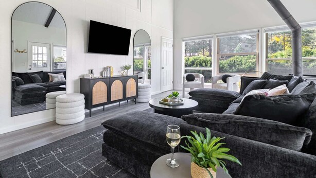 A modern living room with a large flat-screen TV, a black sofa set, a white armchair, and a dining table with a glass of wine. - Golf View Home with Fire Pit & BBQ (Flagstaff)