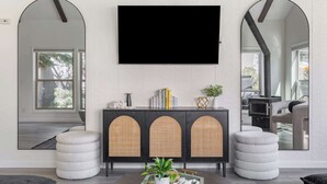 A modern living room with a flat-screen TV mounted above a dark wooden cabinet with woven doors, a round coffee table with decorative items, and a large window offering a view of the outdoors. - Golf View Home with Fire Pit & BBQ (Flagstaff)
