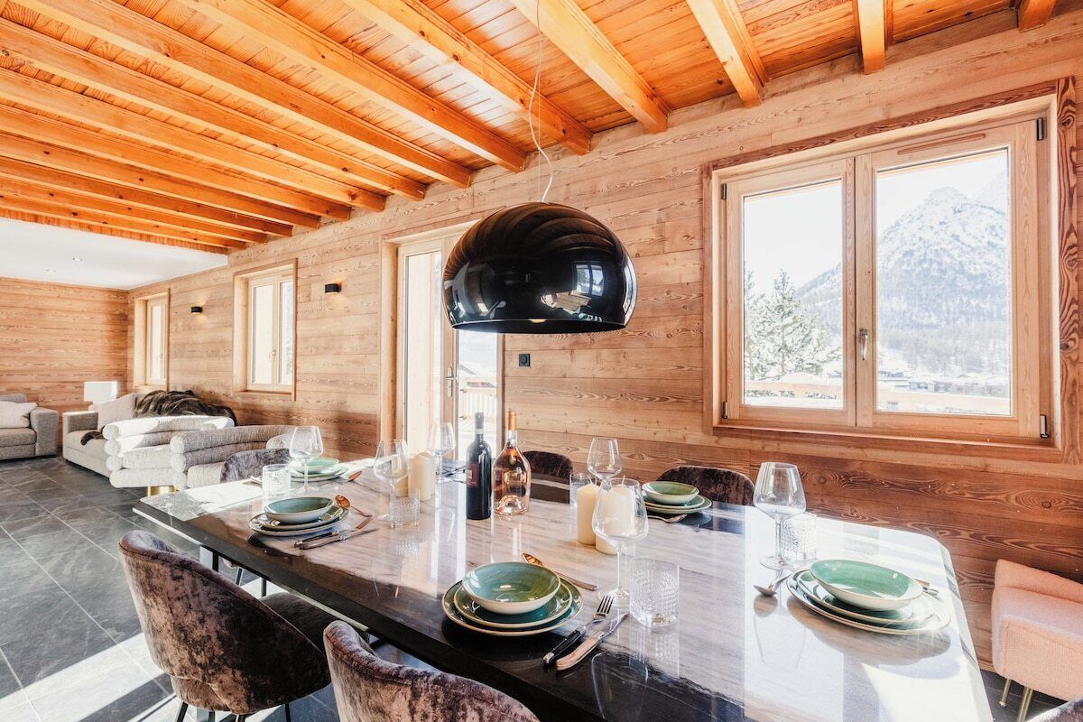 A wooden interior with a dining table set for six, large windows, and a mountain view.