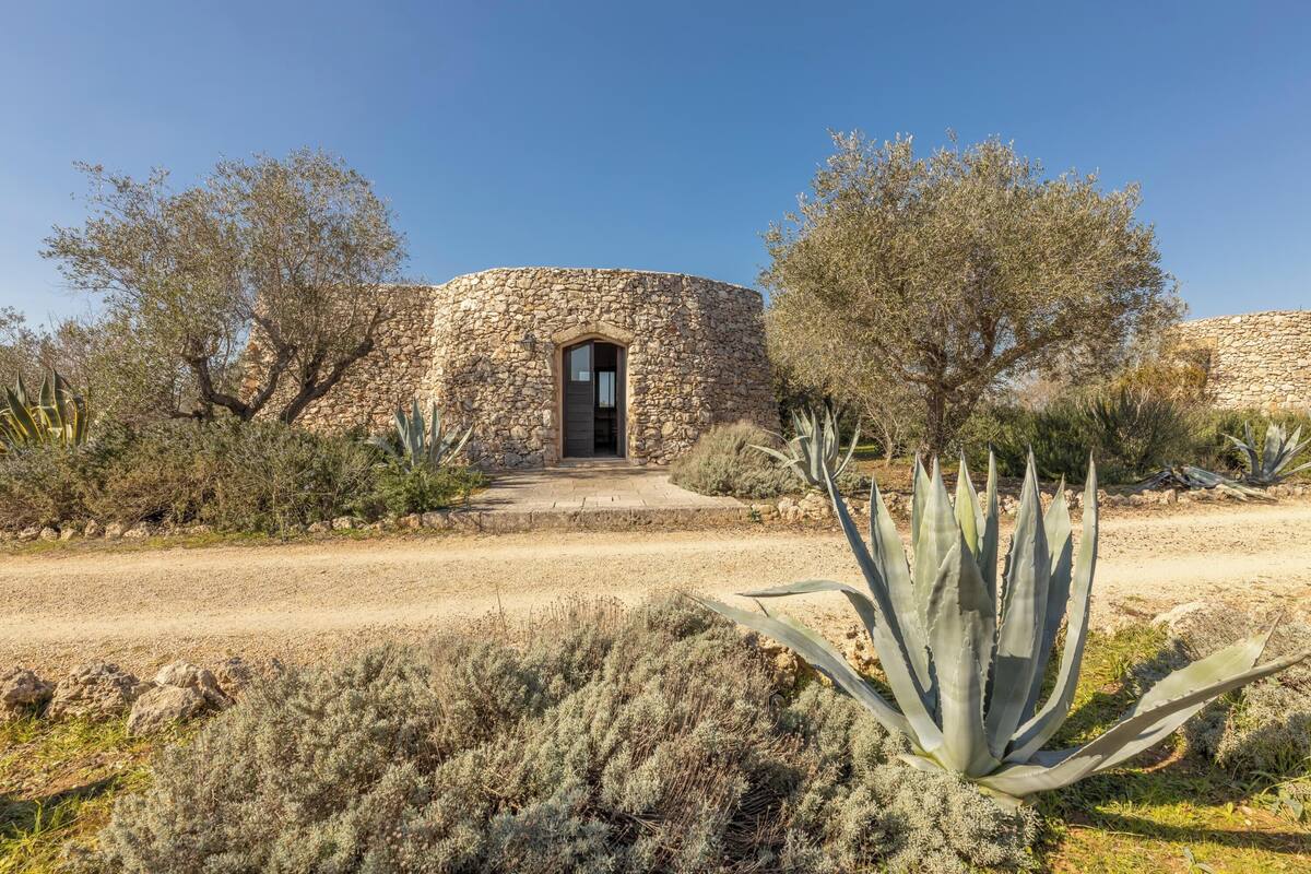 A stone building with a central doorway, surrounded by desert vegetation and trees.