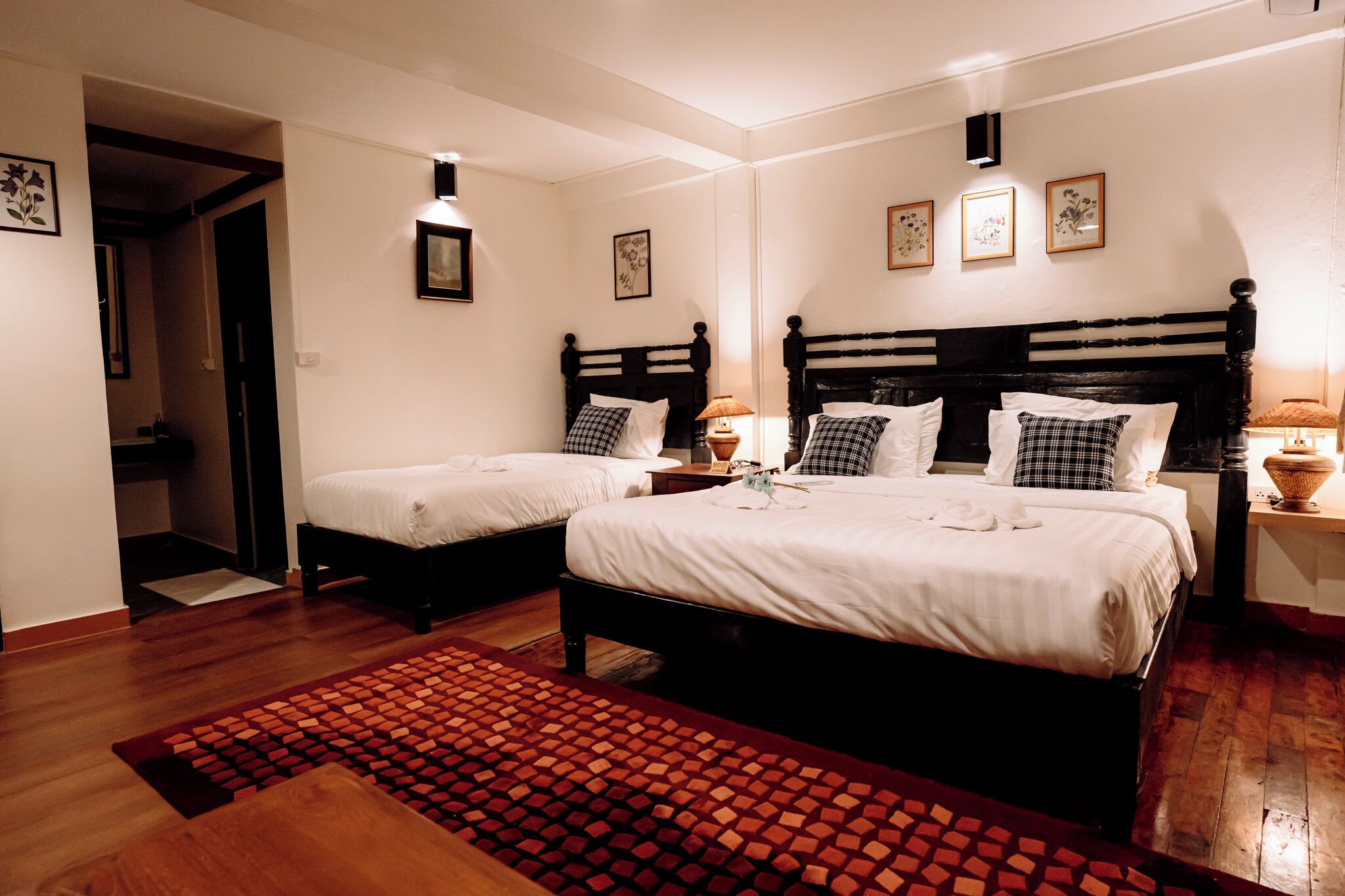 Een hotelkamer met twee bedden, een houten vloer, een rood tapijt met patroon en een kleine tafel met een lamp.