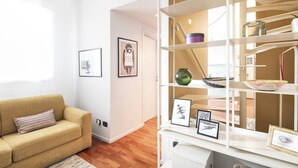 A living room with a beige sofa, a white wall with framed pictures, and a white shelving unit with various decorative items. - Easylife - Cozy two-level unit in Isola (Milano)