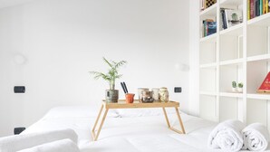 A neatly made bed with white linens, a bedside table with a potted plant, and a bookshelf with books and decorative items. - Easylife - Cozy two-level unit in Isola (Milano)