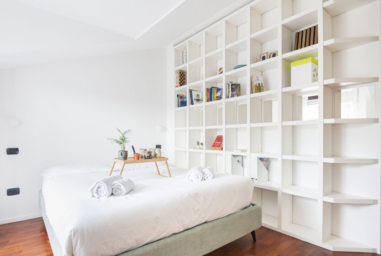 Un dormitorio ordenado con una amplia estantería blanca, una cama con toallas dobladas y una pequeña mesita de madera con una planta.
