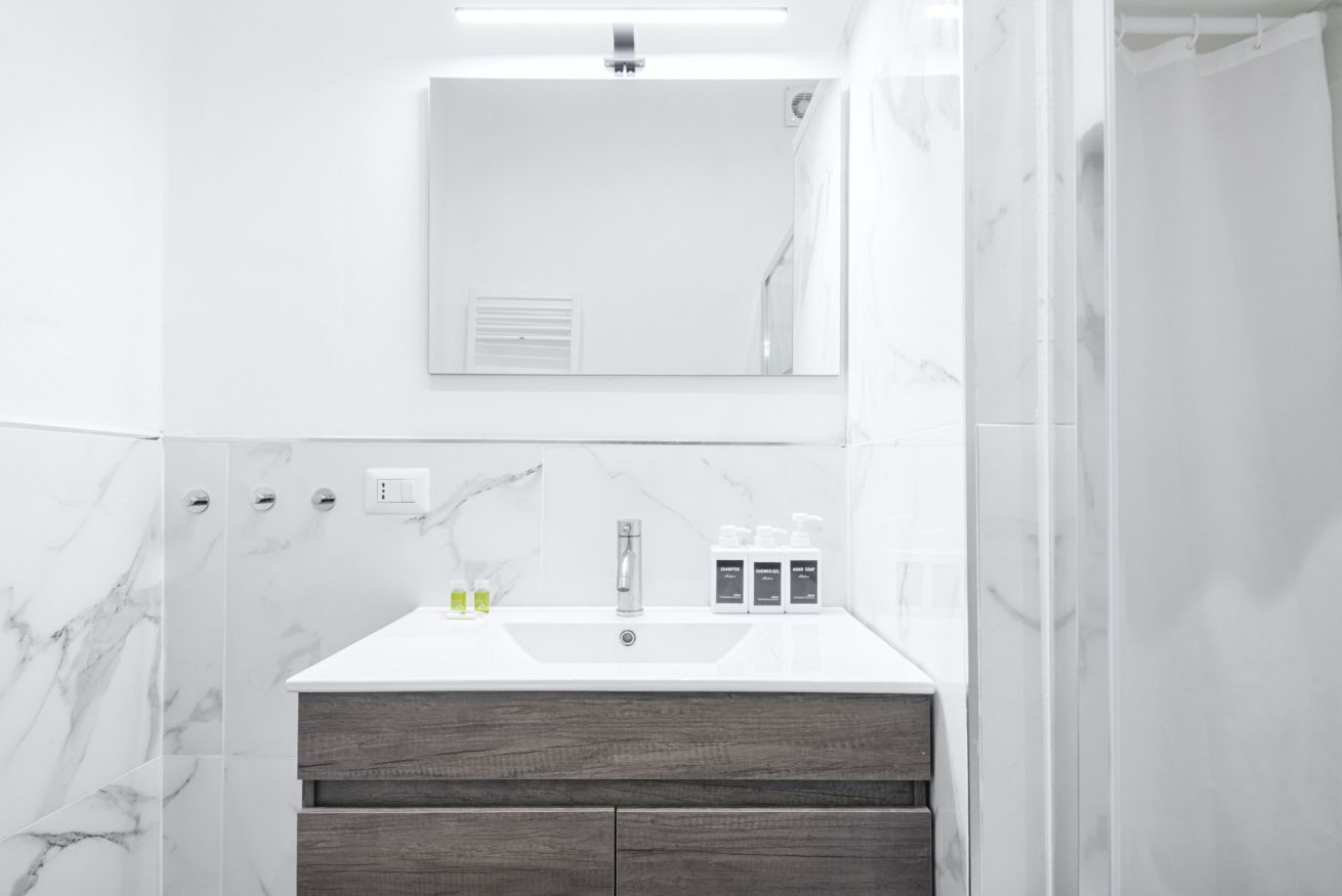 A modern bathroom with a white sink, marble tiles, a wooden vanity, and a mirror above the sink.