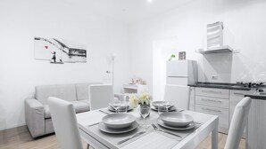Une cuisine moderne et minimaliste, avec des meubles blancs, une table à manger dressée avec de l’assiette et des couverts, et un canapé gris dans l’espace salon.