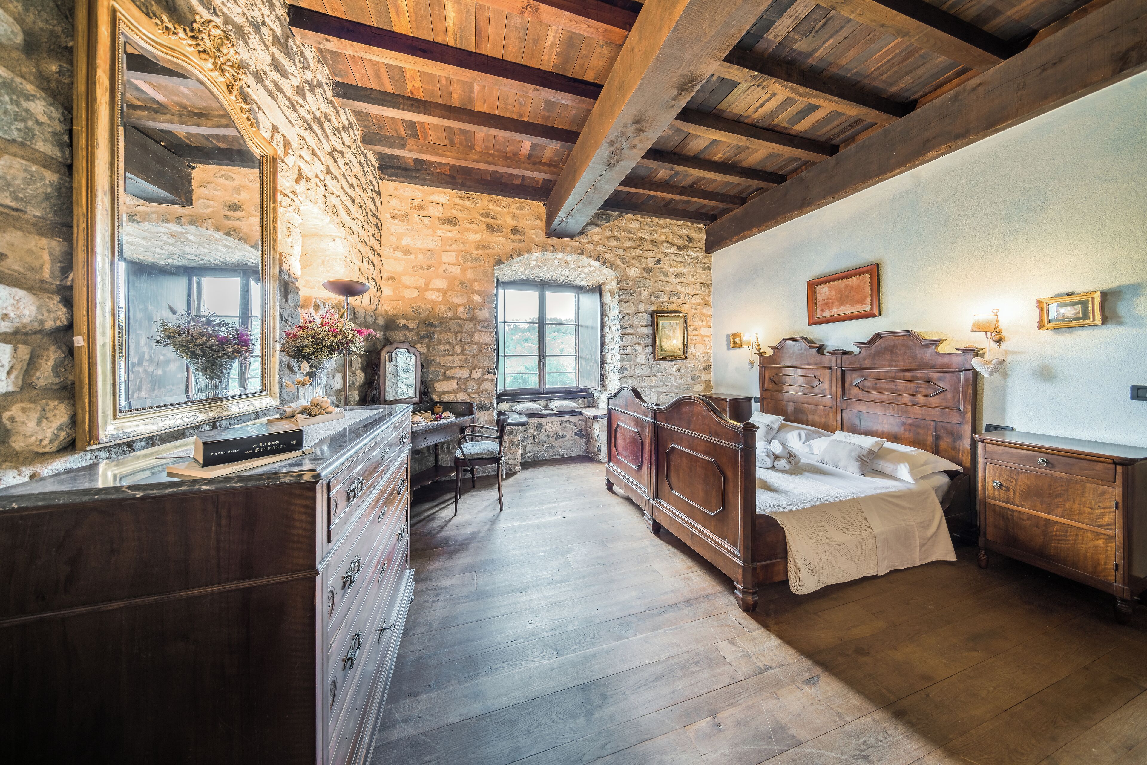 Een landelijke slaapkamer met een stenen muur, houten balken in het plafond, een grote spiegel, een houten bed, een dressoir en een raam met uitzicht.