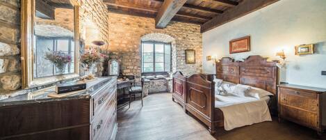 Een landelijke slaapkamer met een stenen muur, houten balken in het plafond, een grote spiegel, een houten bed, een dressoir en een raam met uitzicht.