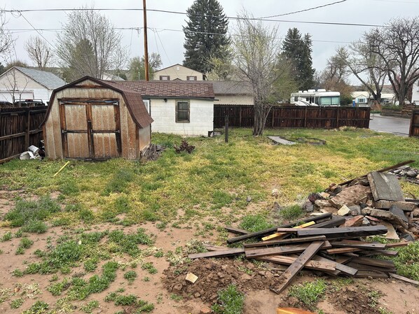 A backyard with a wooden shed, a small house, and a pile of wooden planks.