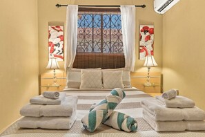 A hotel room with a bed, two bedside lamps, a window with decorative iron bars, and artwork on the walls.