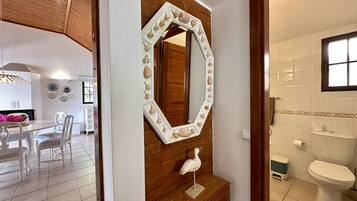 Een kamer met een houten wand, een witte spiegel met schelpen, een witte sculptuur van een vogel, een zithoek met stoelen en een badkamer met toilet en wastafel.
