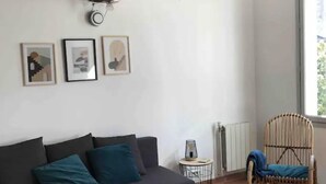 Una sala de estar con un sofá oscuro, una mesa redonda de centro y un sillón de mimbre. Hay cuadros enmarcados en la pared y una ventana que proporciona luz natural.